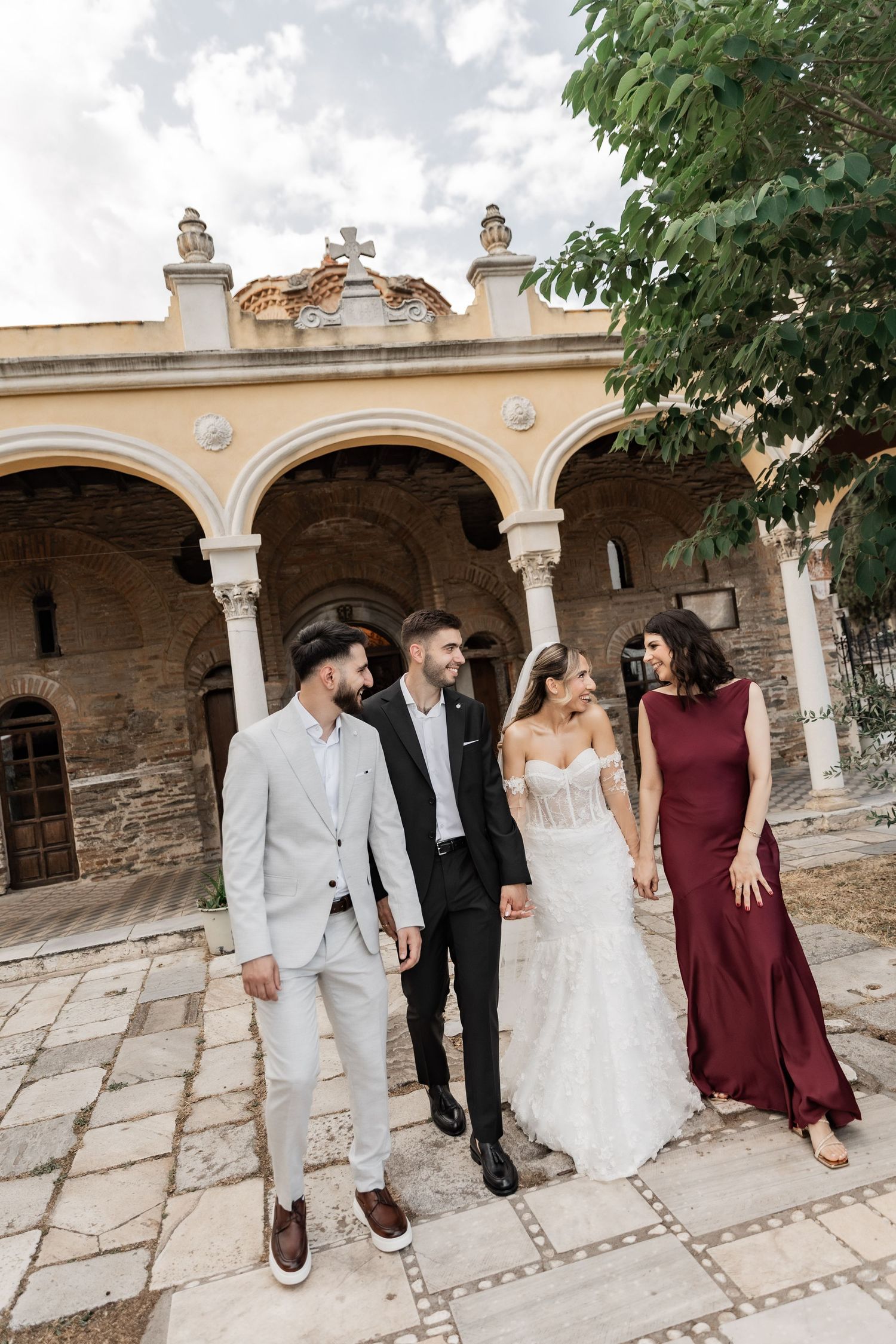 Bride and groom with wedding guests posing outside a historic European church with arched colonnade.