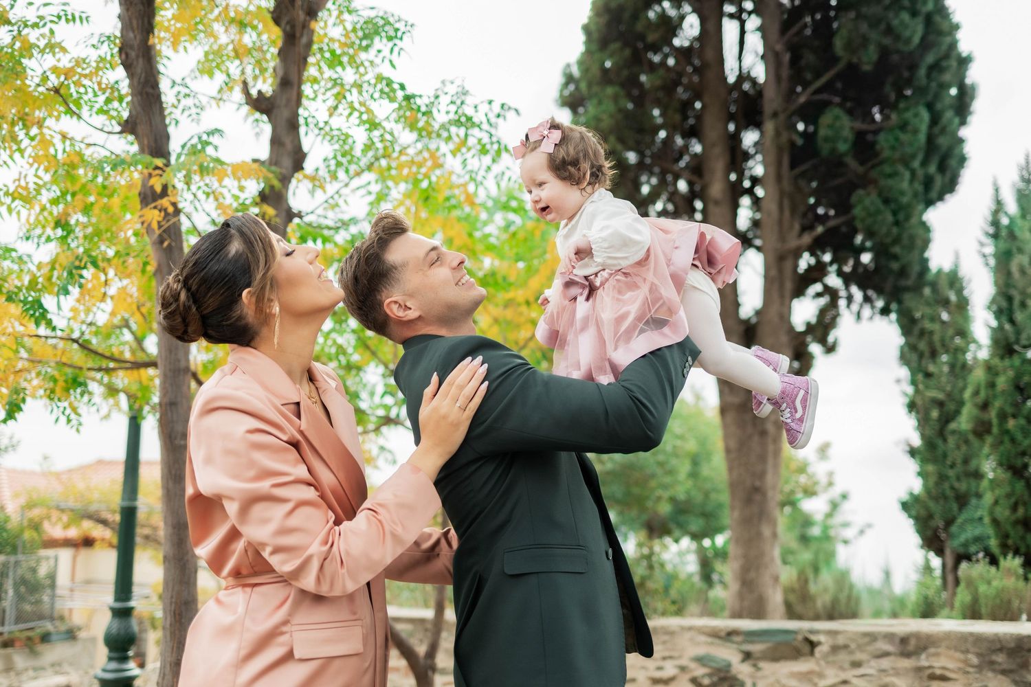 A couple shares a romantic moment in an outdoor garden setting with autumn trees and cypress in the background.