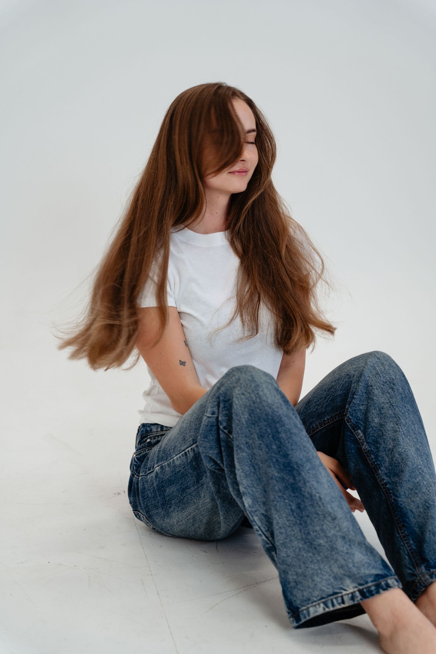Young woman with flowing brown hair sits on white floor wearing white t-shirt and blue jeans in studio setting.