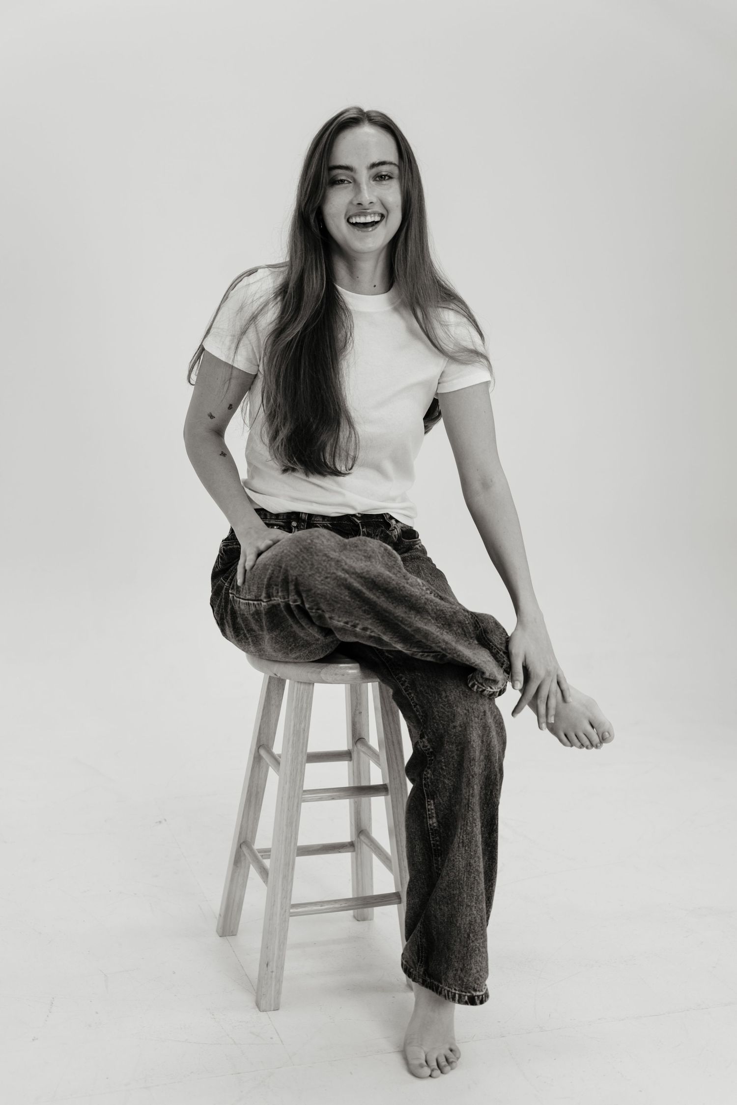 Black and white photo of a smiling young woman sitting on a stool, wearing a white t-shirt and jeans, barefoot.