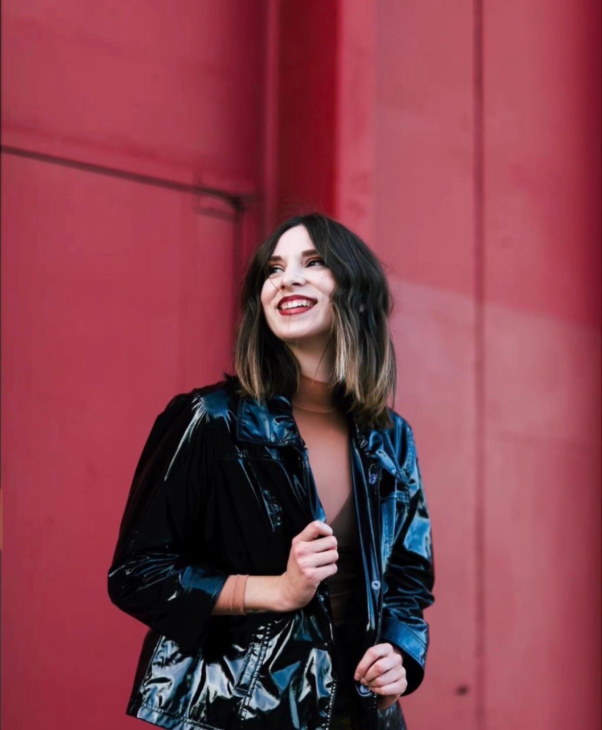 Smiling woman with ombre bob wearing shiny black jacket poses against vibrant red wall.