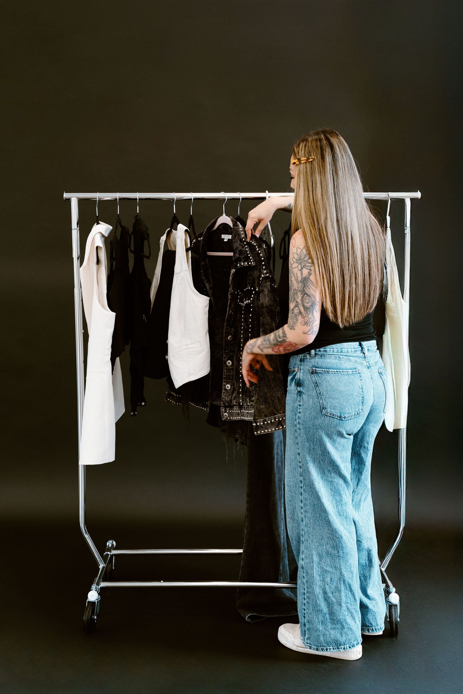 Tattooed woman with long blonde hair browsing black and white clothing on a rack, wearing jeans and black top.