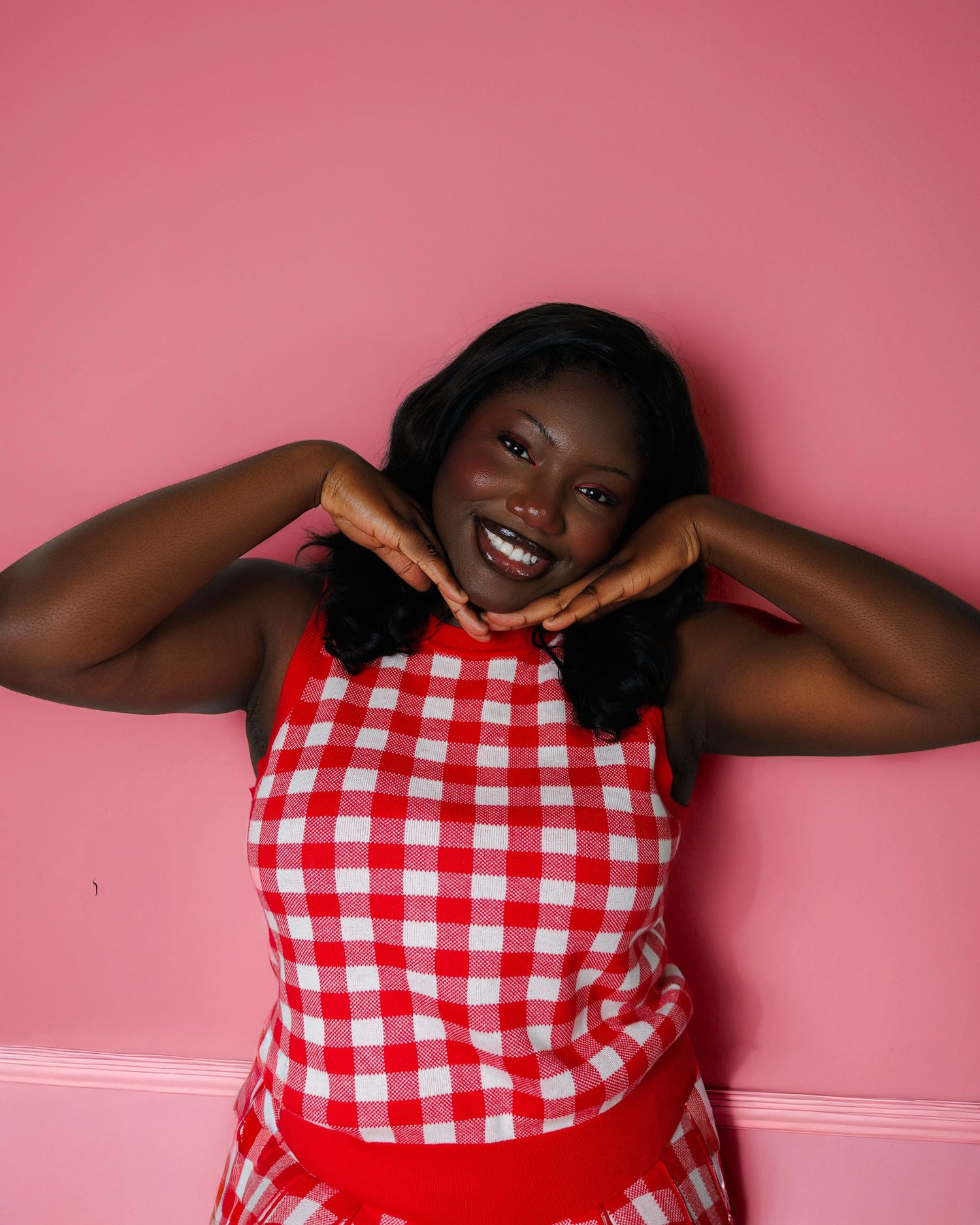 Smiling woman in red gingham dress posing against pink wall with hands framing her face.