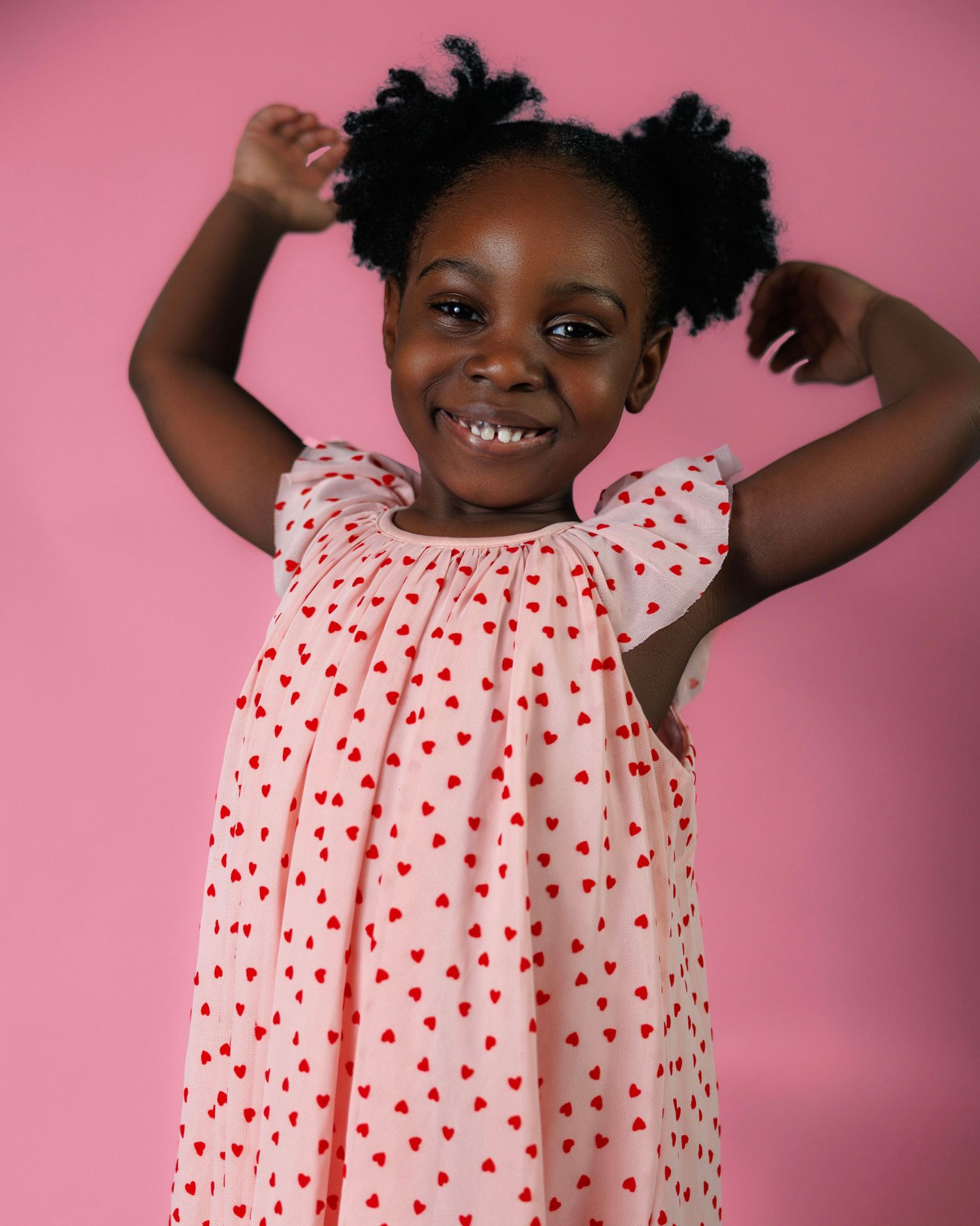 A joyful young girl in a pink heart-print dress poses with arms raised, smiling brightly against a pink background.