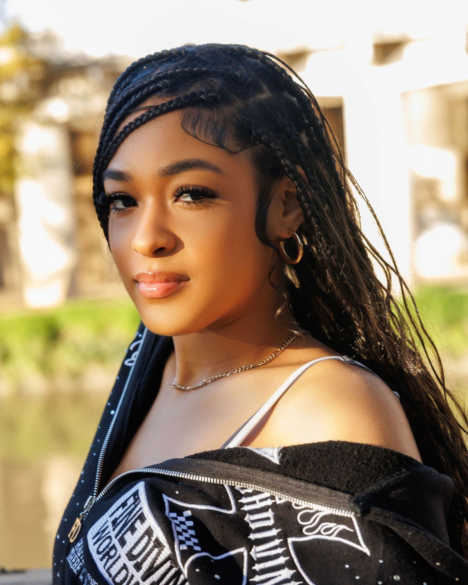 Young woman with long box braids wearing gold hoop earrings, a necklace, and an off-shoulder graphic jacket outdoors.