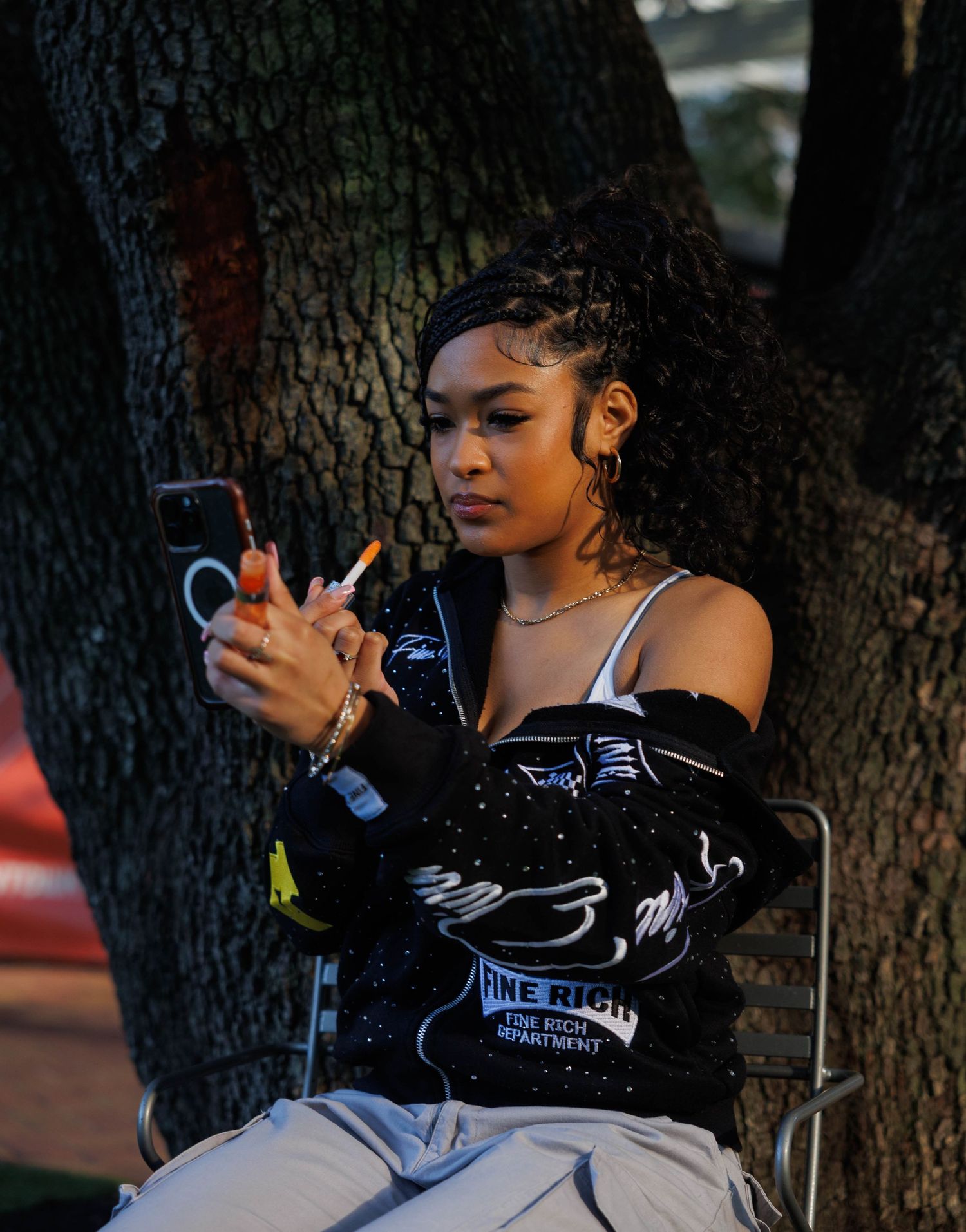 Person sitting outdoors against a tree trunk using a mobile phone in casual attire.