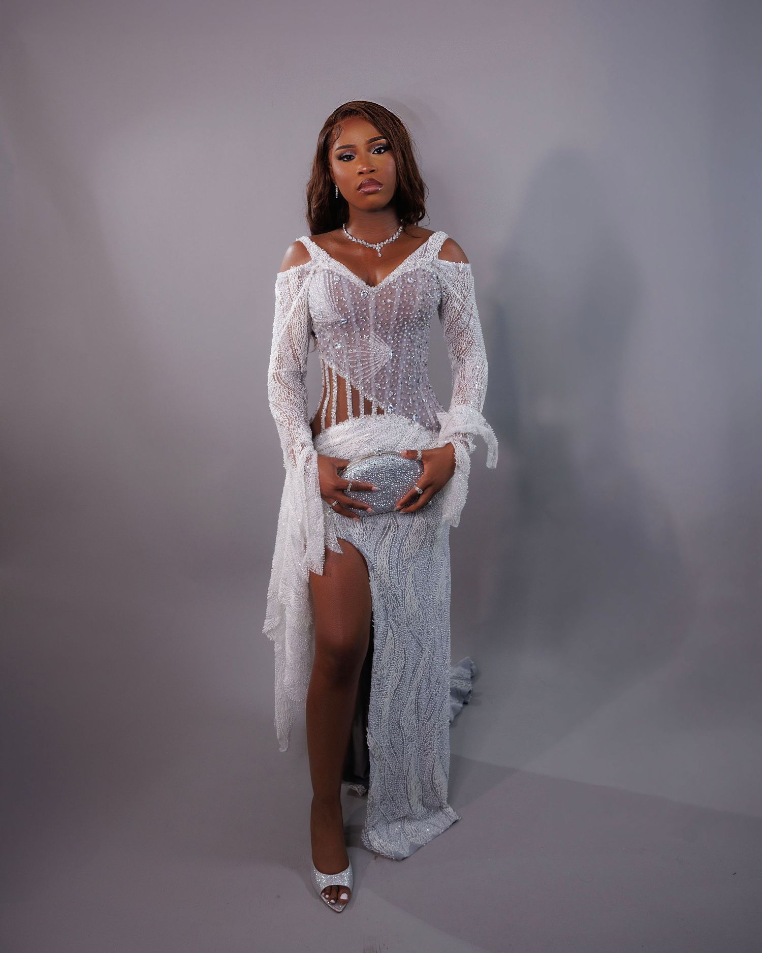 Woman in white lace sheer gown with long sleeves, high slit, and silver accessories poses elegantly.