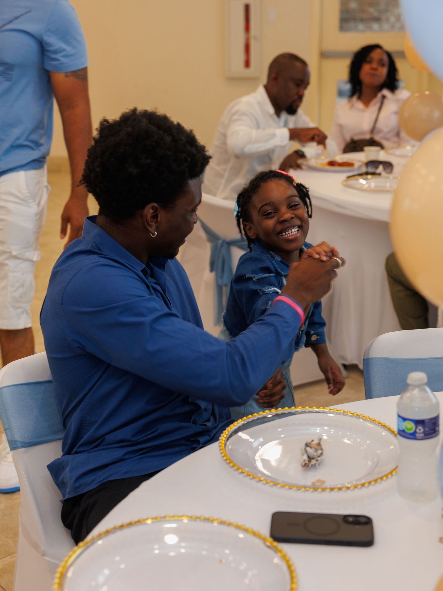 A woman and young girl in blue outfits share a joyful moment at an elegant event table with gold-rimmed plates.