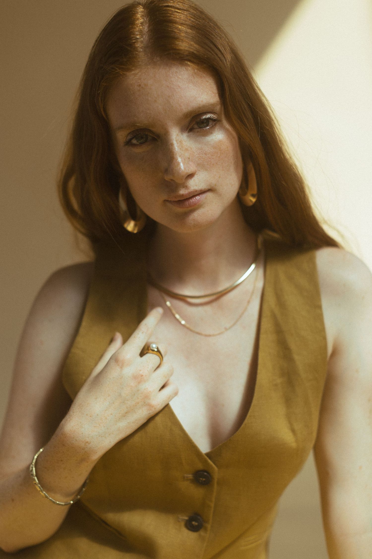 Person wearing a mustard yellow vest and delicate jewelry poses in warm sunlit portrait with natural styling.