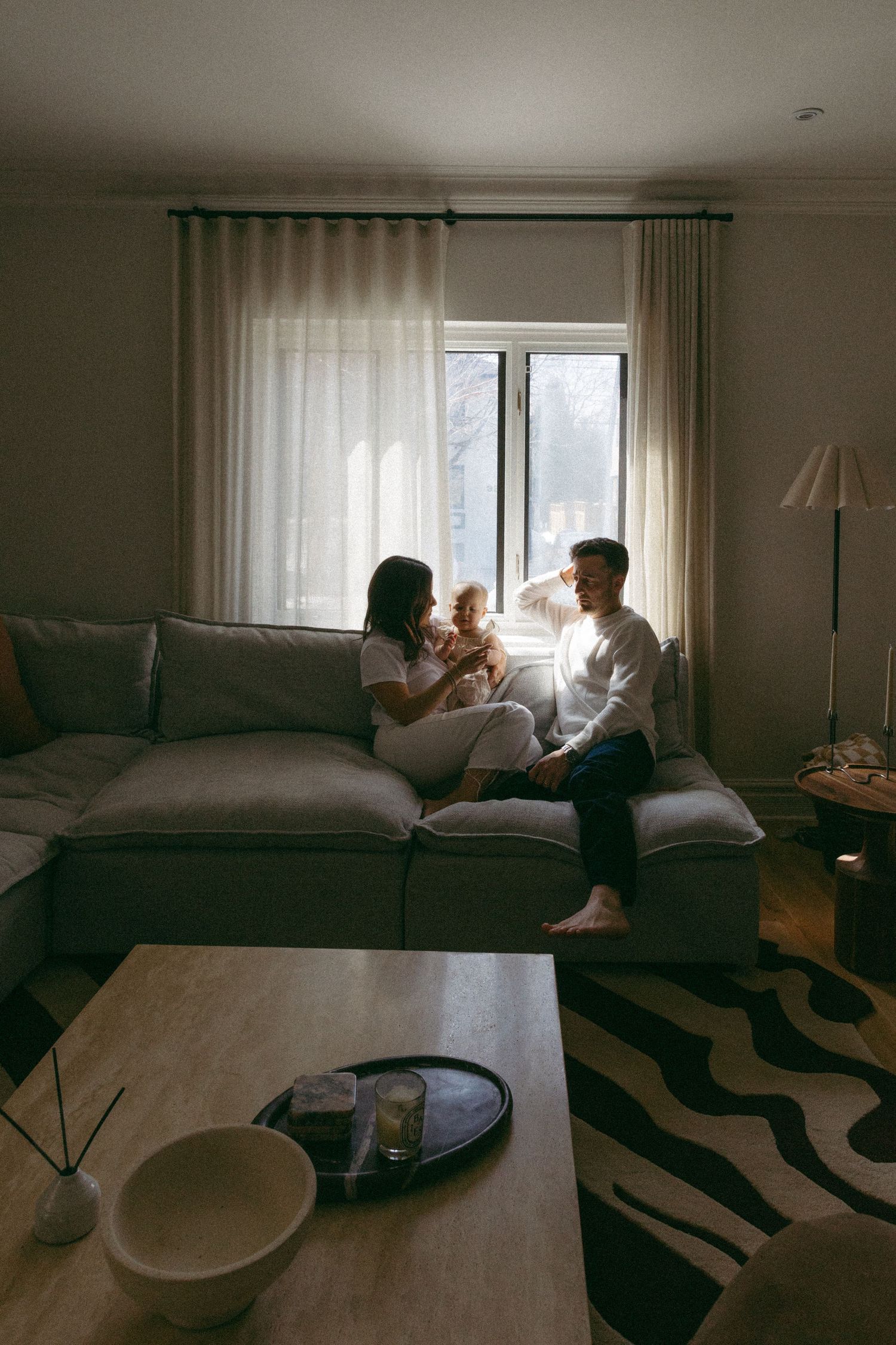 People sitting together on a gray couch in a softly lit living room with white curtains and wooden coffee table.