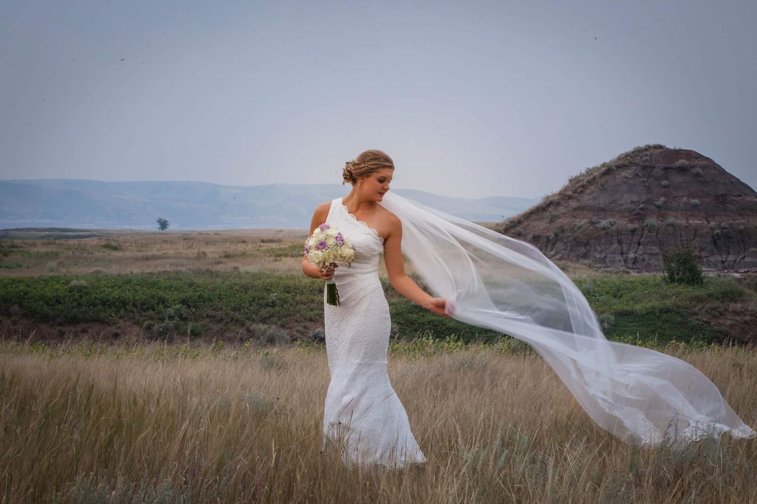Bride in white gown stands in a field as her veil flows dramatically in the wind with mountains in background.