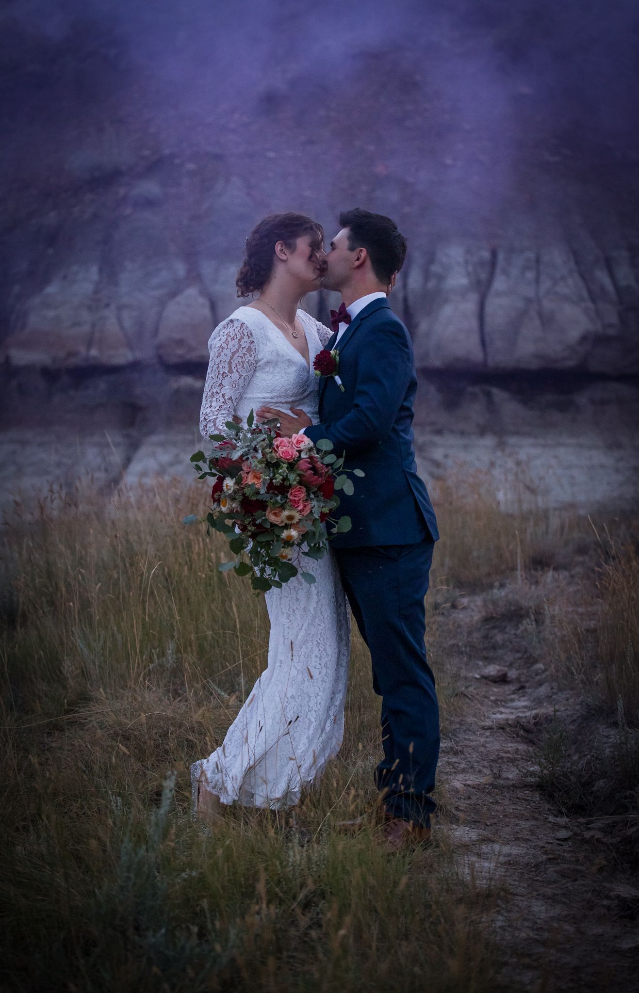 A couple shares a romantic kiss in a field at dusk wearing a white lace wedding dress and navy suit.