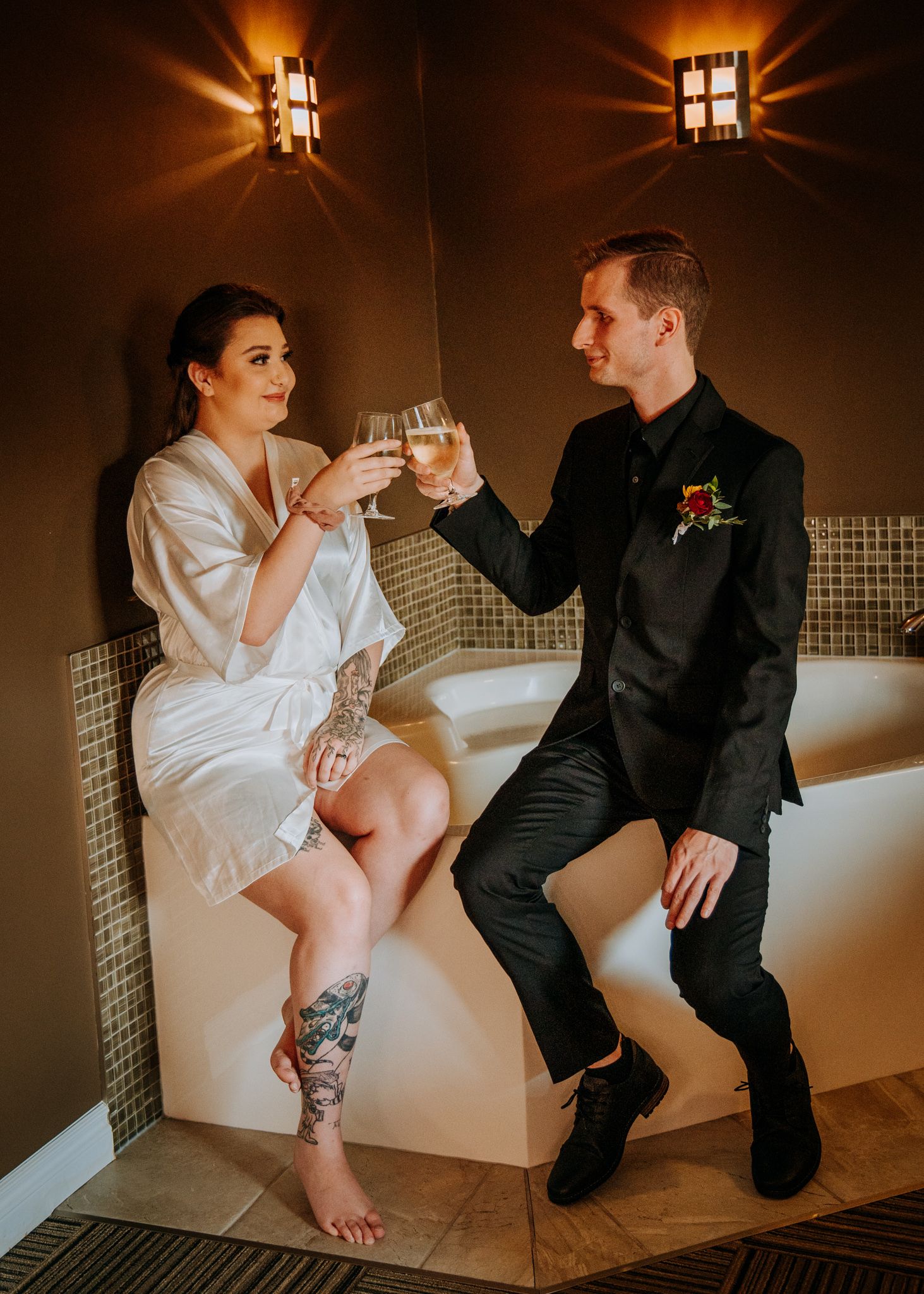 A couple in wedding attire sits on a bathroom floor showing off their matching leg tattoos in warm lighting.