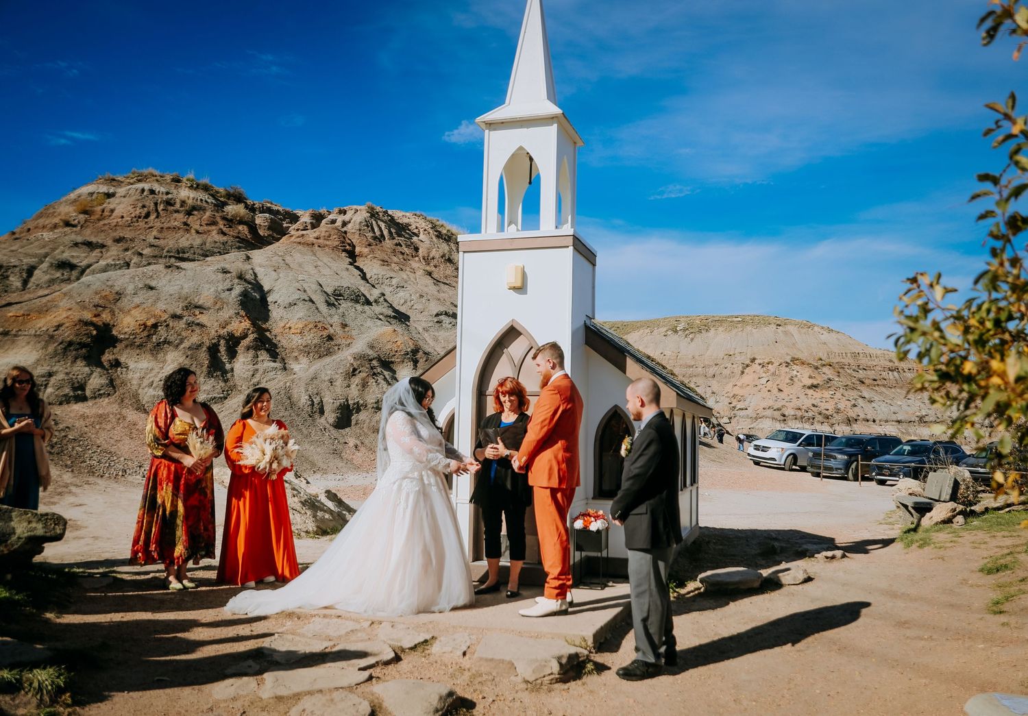 Wedding ceremony takes place outside a white chapel with steeple in a desert mountain landscape.