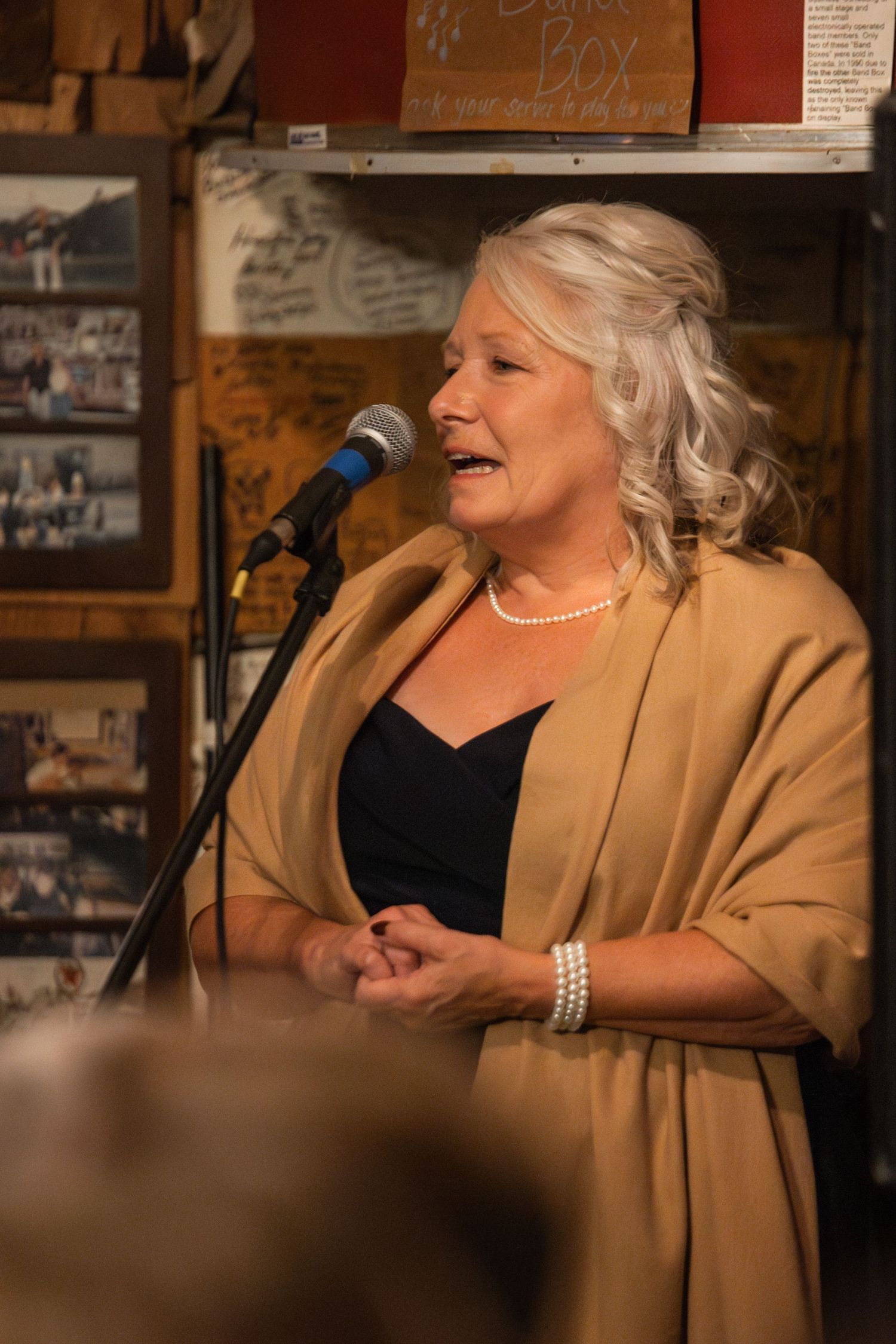 A performer in a beige jacket and black top sings into a microphone at an intimate venue.