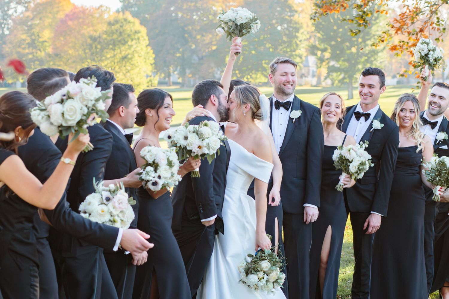 Wedding party in black suits and dresses celebrates with white bouquets and autumn foliage backdrop.