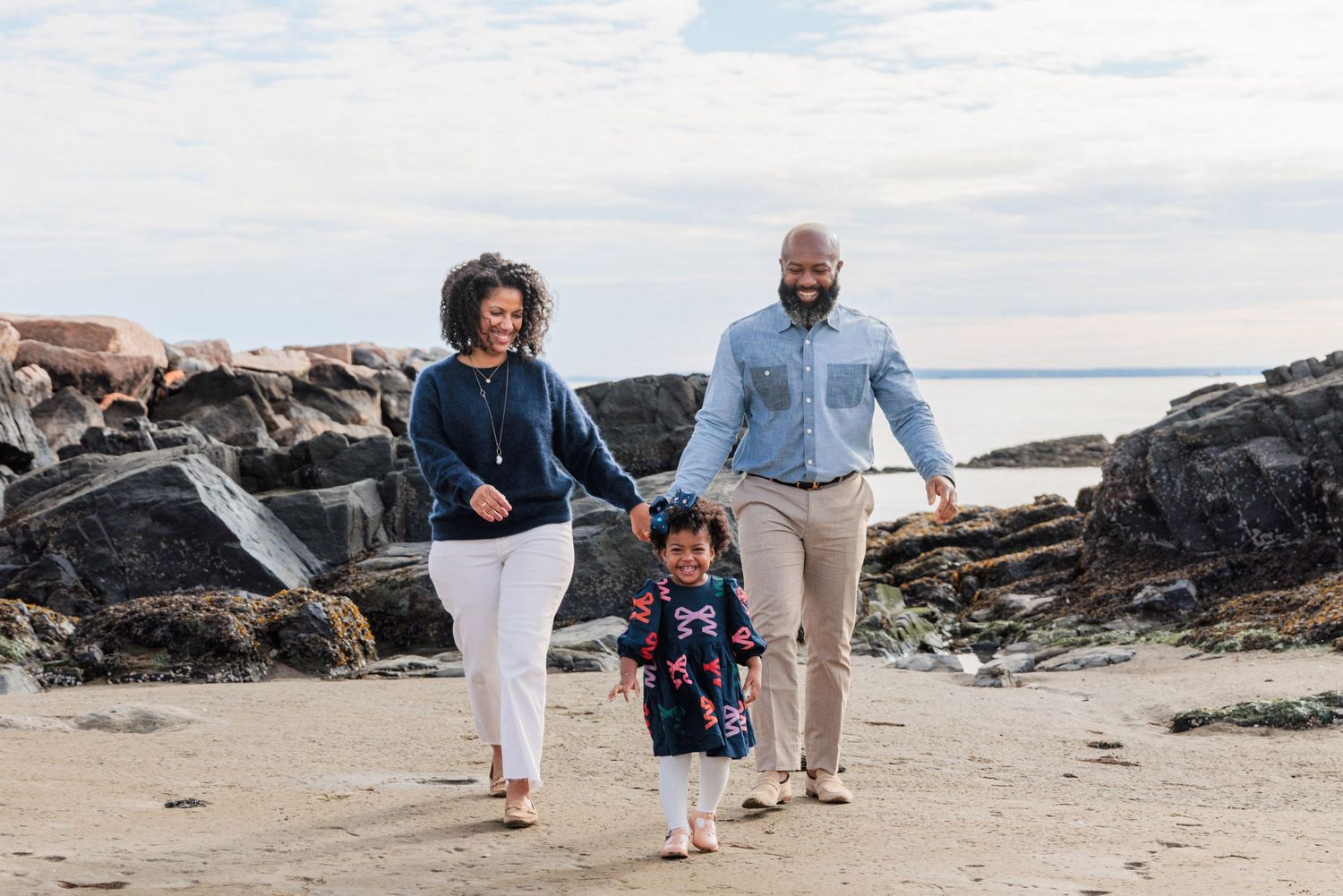 Family walking together on Rye Beach. Behind them are the rocks and the Long Island Sound. The daughter is slightly in front of her parents.