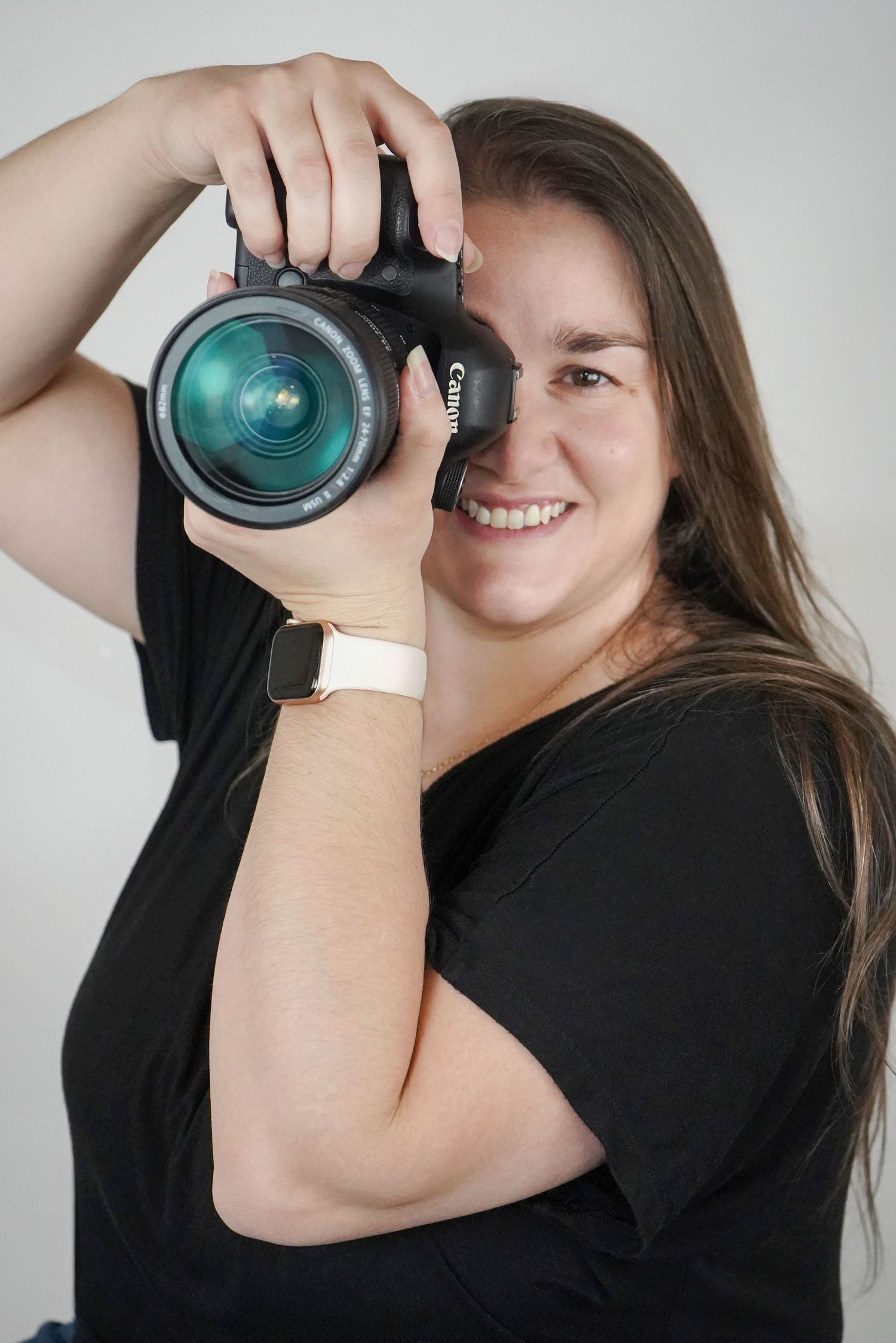 A photographer wearing a black shirt holds up a professional DSLR camera with a large lens against a light gray background.