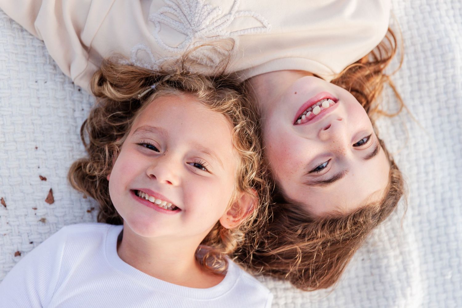 Closeup of two sisters laying on a white blanket. One is upside down to the other and their heads are nestled together. Both are smiling.