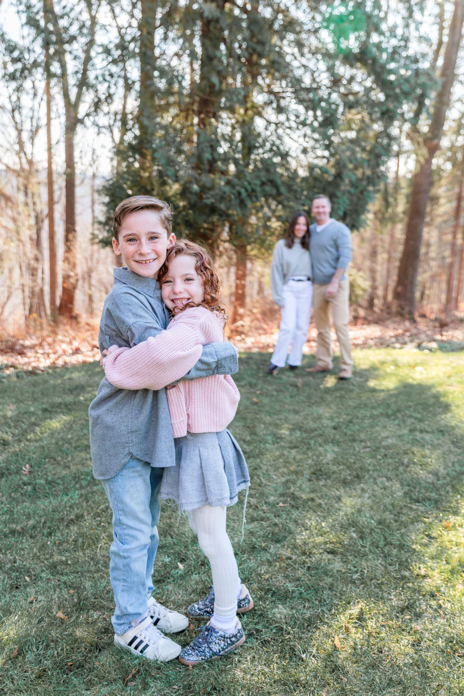 Two kids stand in the foreground of the photo hugging. Behind them, mom and dad are smiling and blurred out.