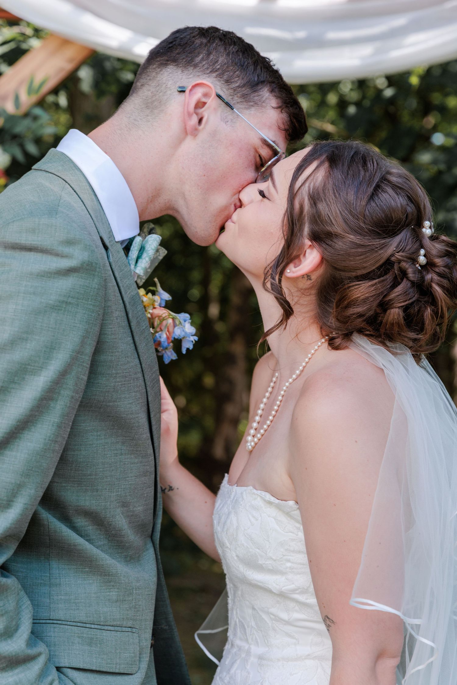 Bride and groom share a staged first kiss at outdoor wedding ceremony, bride in white dress with pearl necklace.