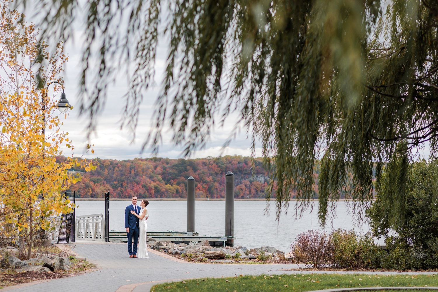 A couple embraces under a weeping willow tree along the Hudson River in autumn.