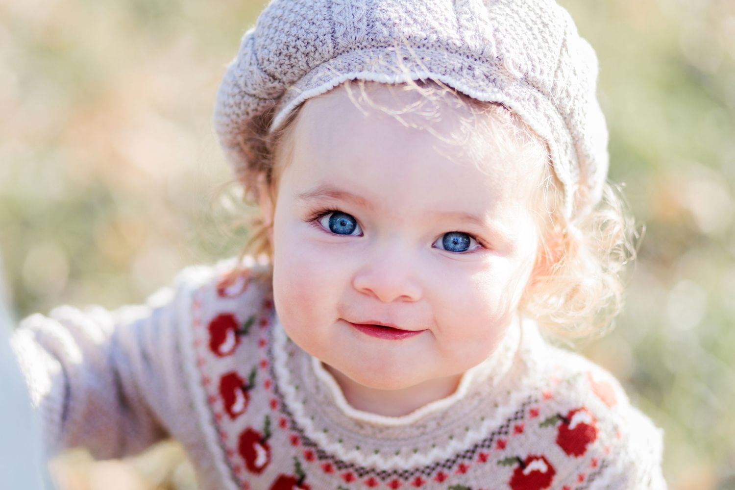 A baby wearing a floral dress and white headband sits outdoors in soft natural lighting.