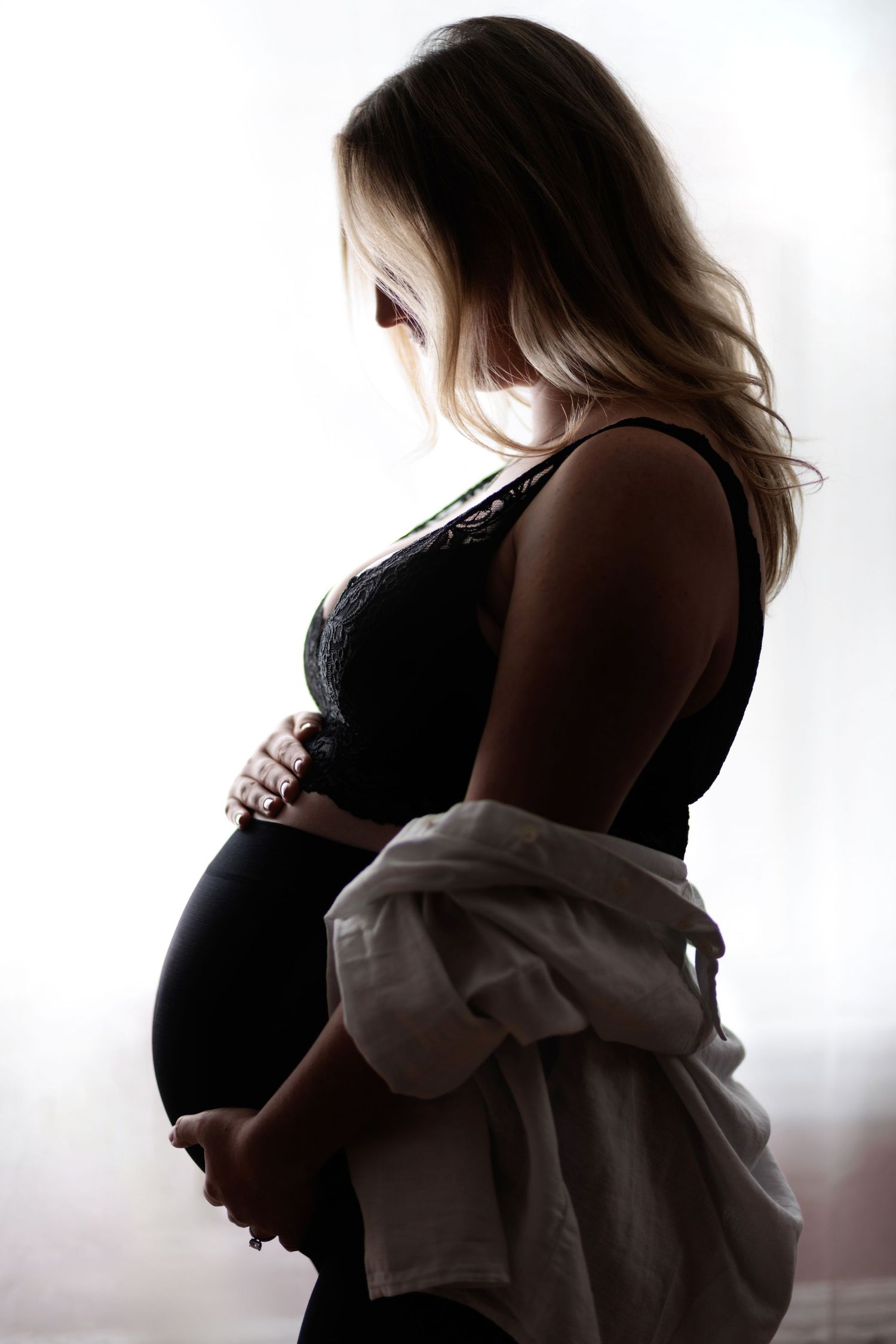 Pregnant woman silhouetted. She's wearing black clothes with a white button down draped over her arms. She's looking and touching her belly.