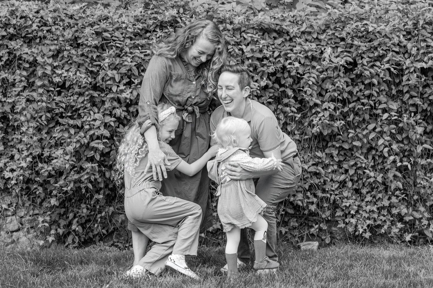 Mom and dad standing together in front of a wall full of Ivy. Dad is lifting youngest girl off the floor and older girl is hugging mom.