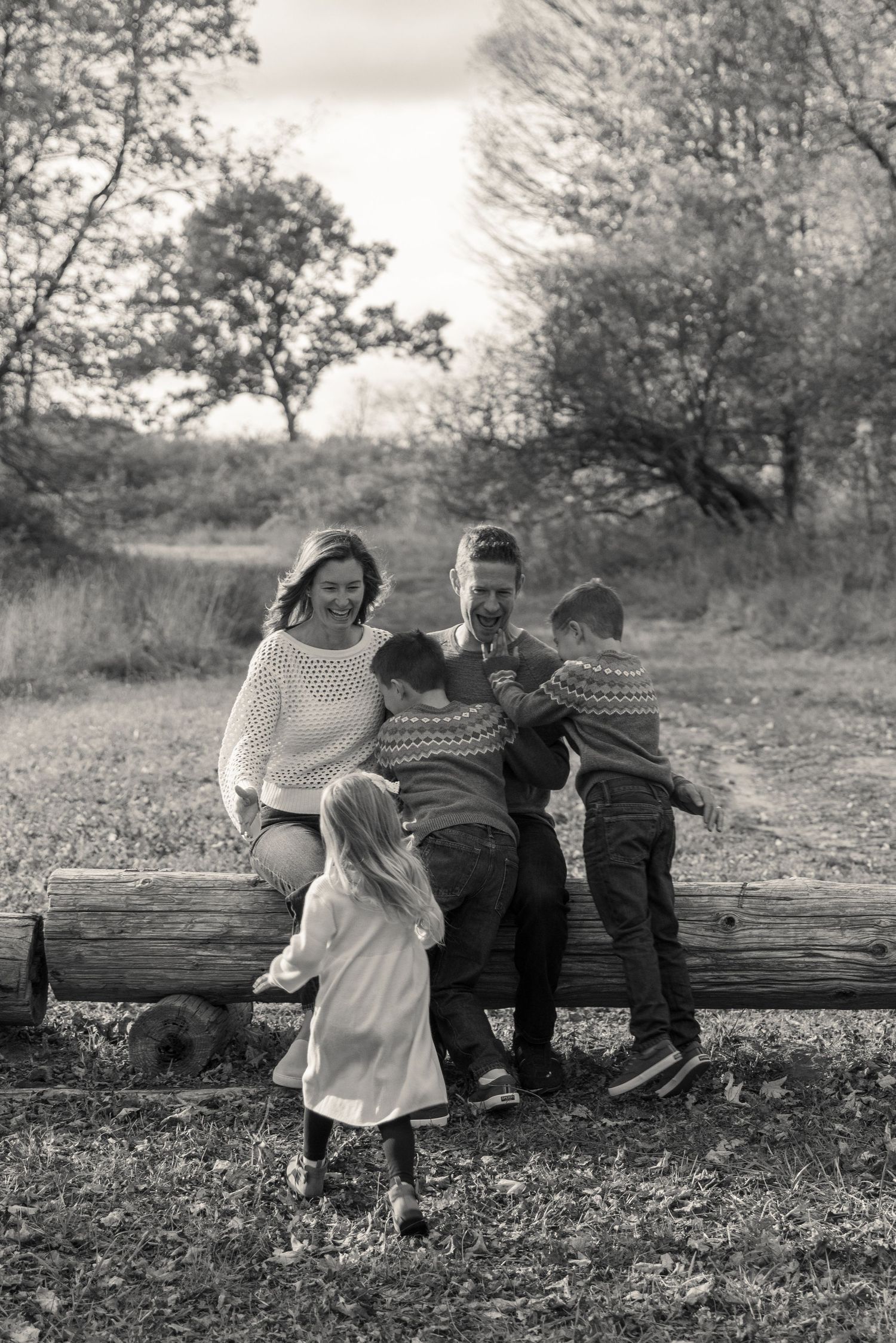 A black and white photo of a Mom and Dad sitting together on a rustic log in a pastoral outdoor setting. Their 3 kids are running to them.