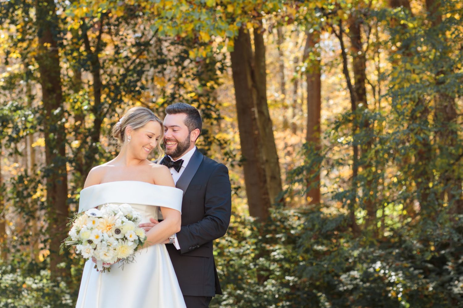 A couple shares a romantic moment in a sunlit autumn forest while dressed in wedding attire.