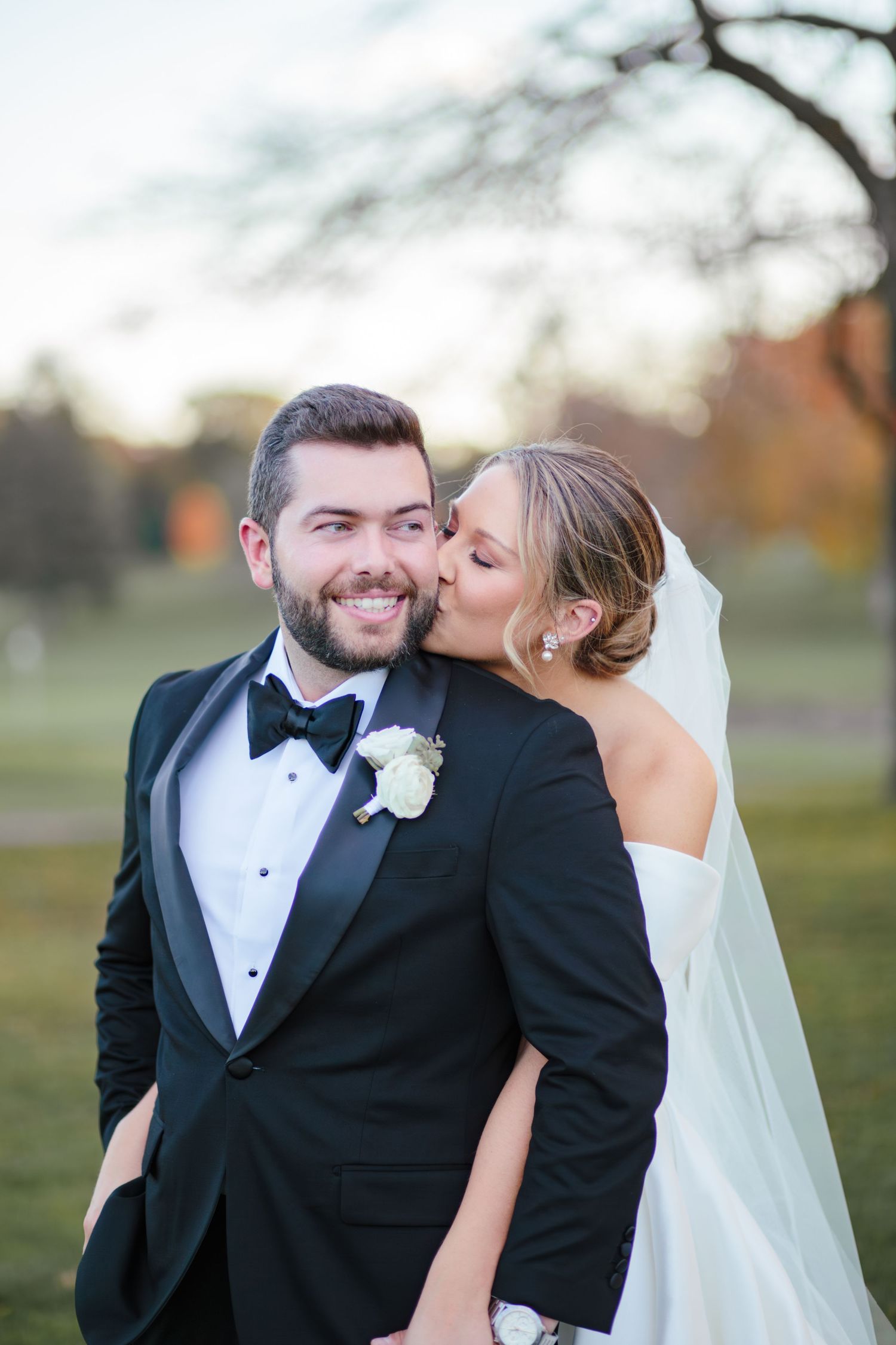 A romantic wedding moment outdoors with the bride wearing a veil and the groom in a black tuxedo with bowtie.