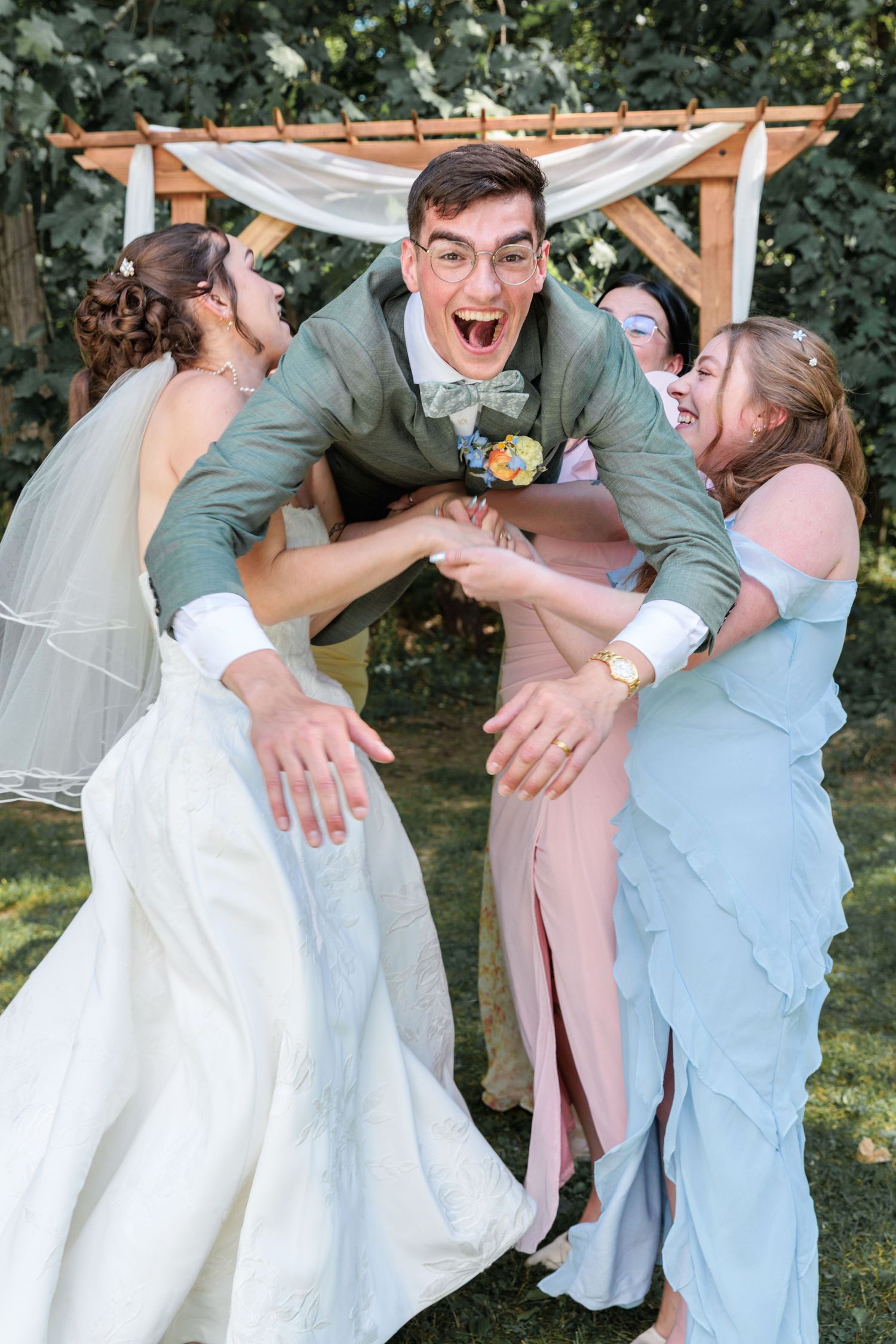 Excited groom in sage suit being thrown in the air by bride and bridesmaids at outdoor wedding ceremony altar.