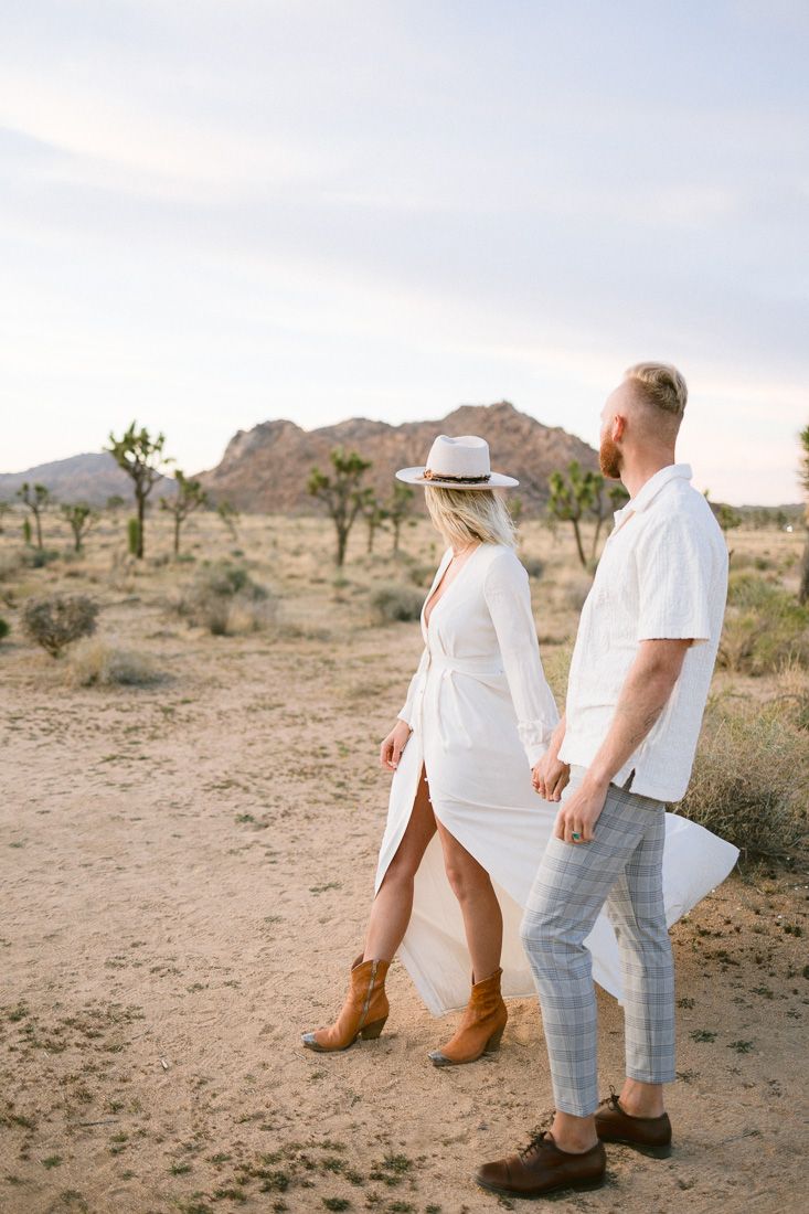 Couple walking hand in hand through Joshua Tree desert landscape at sunset.