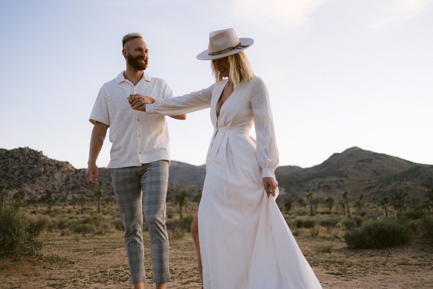 A couple in white outfits share a playful moment in front of desert mountains during golden hour.