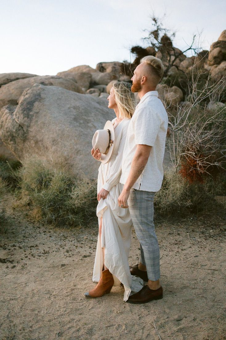 Couple in white clothing standing together against large desert boulders at sunset.