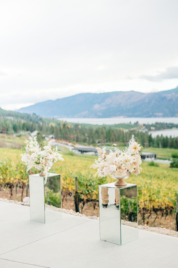 Modern glass pedestal vases with white floral arrangements against mountain vineyard backdrop.