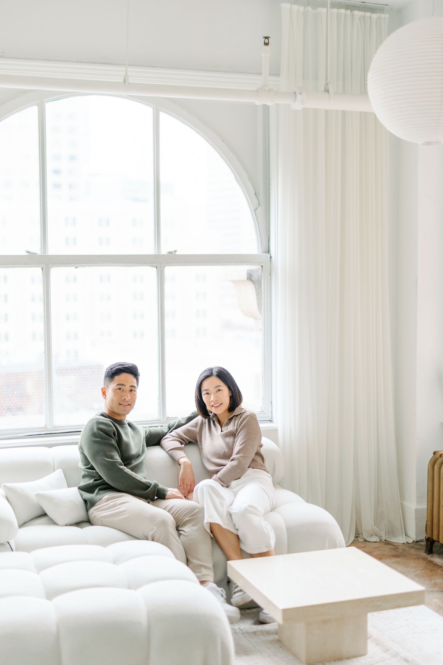 Asian couple sitting together in a cozy white bedroom decorated with minimal modern furniture and natural light.