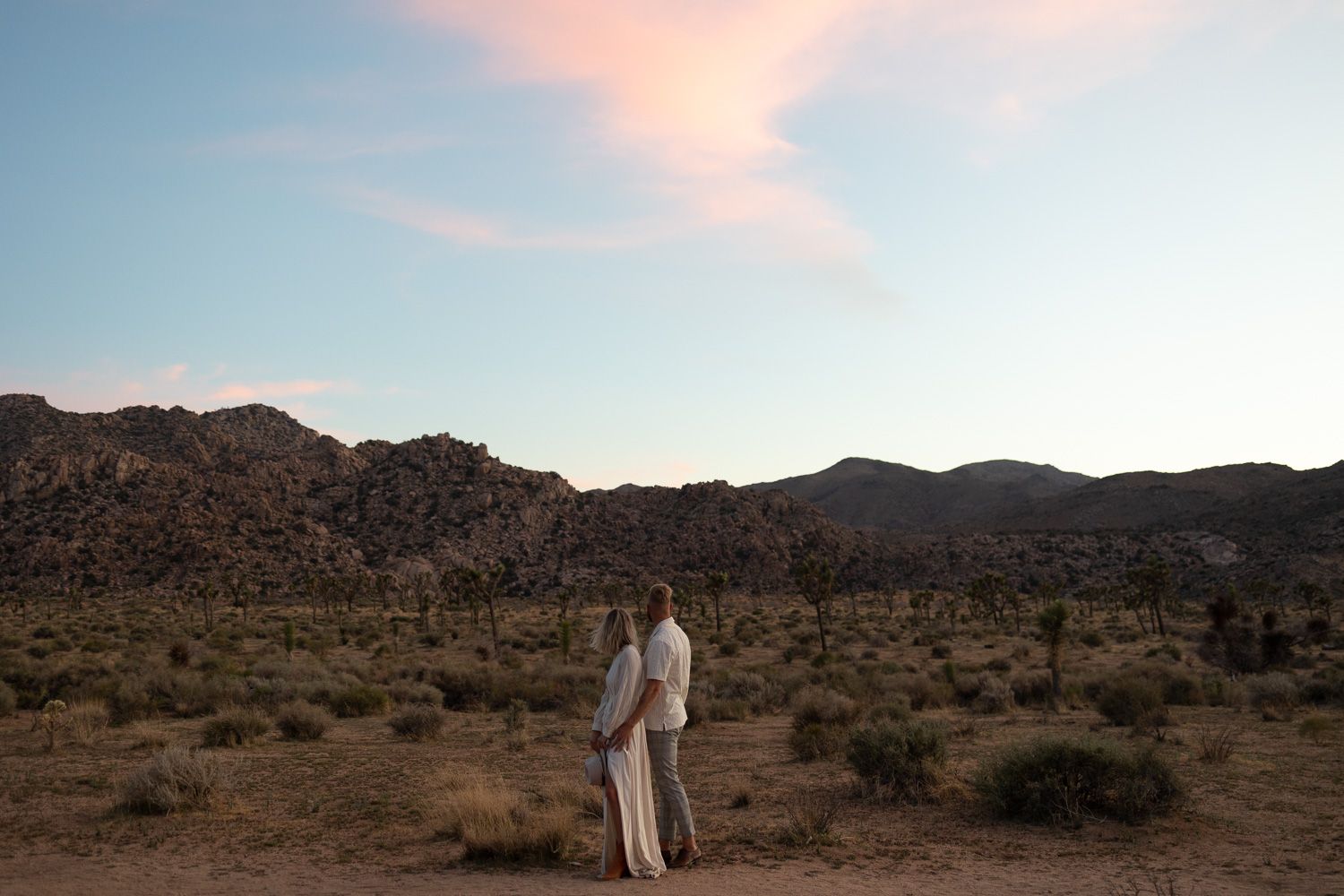 Two people in white stand together against a desert mountain landscape at dusk.