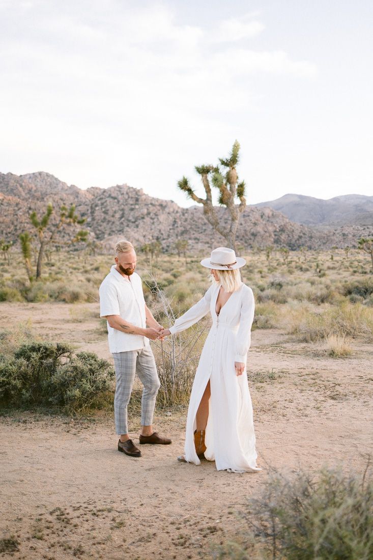 A couple in white outfits holds hands in a desert landscape with Joshua trees and mountains in the background.