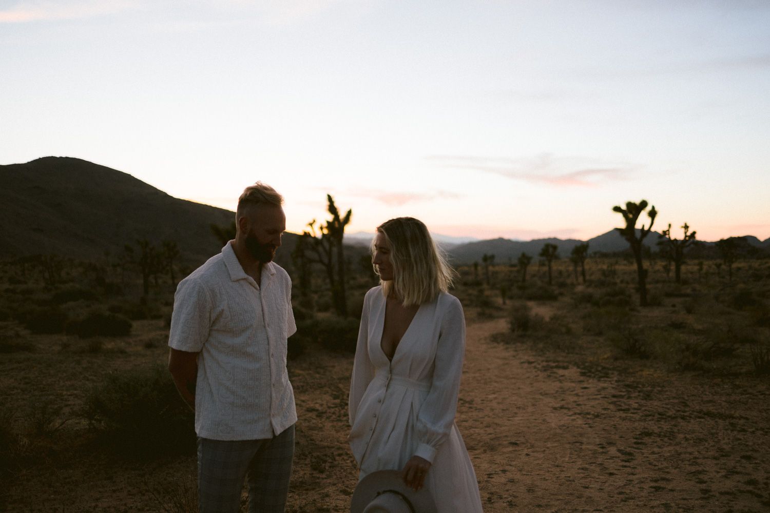 A couple shares an intimate moment on a desert trail with mountains and Joshua trees in the background.