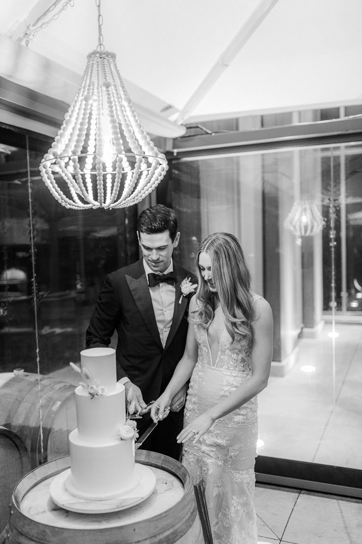 A couple cuts their elegant white wedding cake under a crystal chandelier.