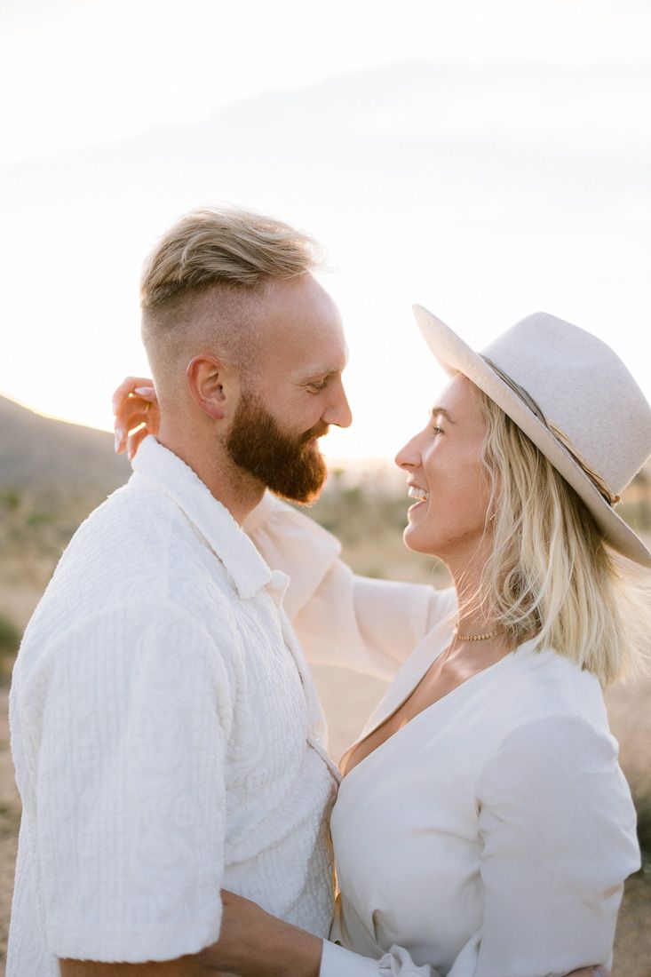A joyful couple shares a close moment in white outfits against a desert landscape.