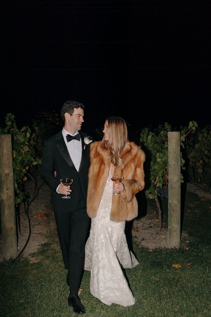 Couple in formal attire sharing wine while standing in a vineyard at night.