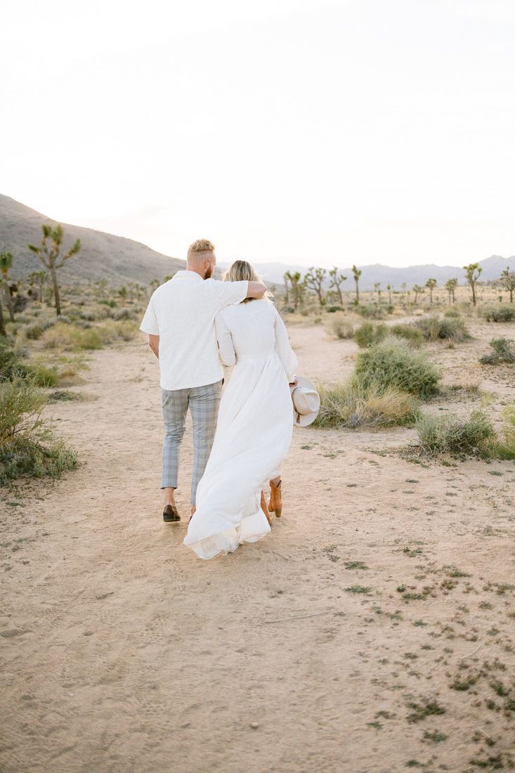 A couple in coordinated white outfits walks through a desert setting with mountains in the background.