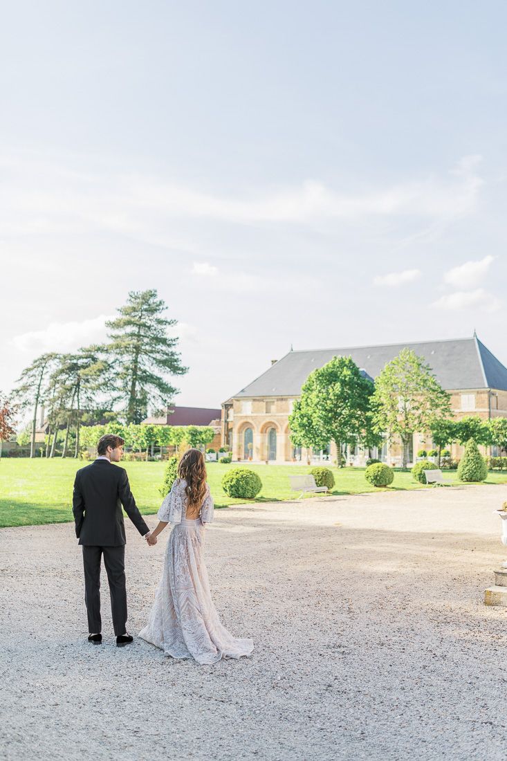 Couple holding hands while walking towards an elegant French chateau on a bright summer day.