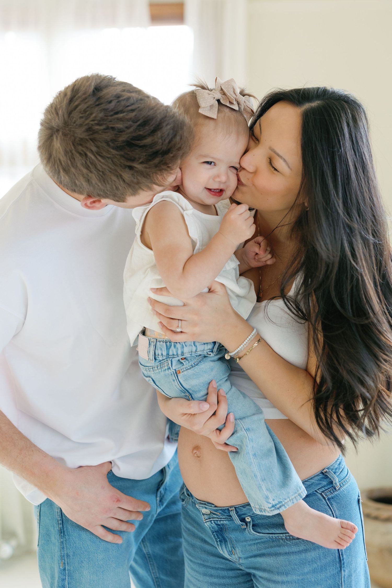 Happy family with parents kissing their smiling baby girl wearing a bow, all dressed in white tops and jeans.