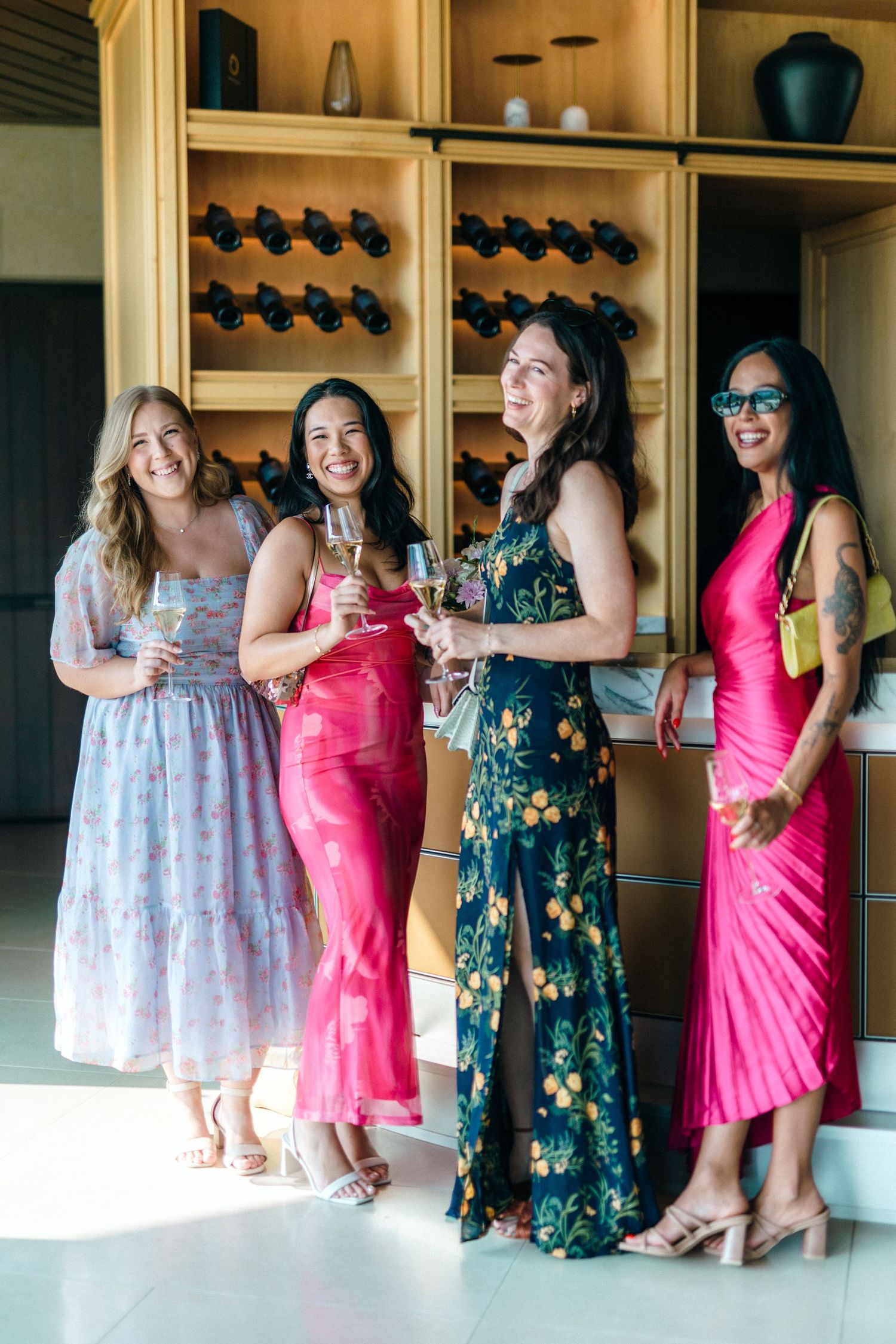 Four women in colorful formal dresses posing together at an elegant wine room gathering.