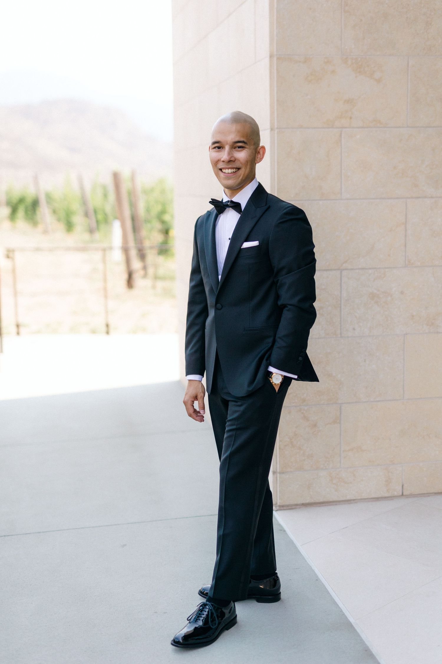 A formal portrait of someone in a tuxedo standing against a light stone wall with mountains in background.