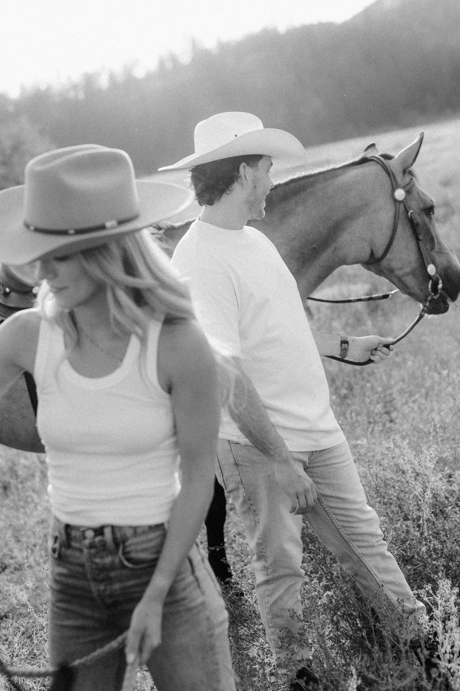 Black and white photo of people in western wear tending to horses in a wild meadow.